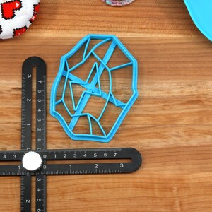 May include: A blue plastic cookie cutter shaped like a geometric design with a web-like pattern. The cutter is on a wooden surface with a ruler.