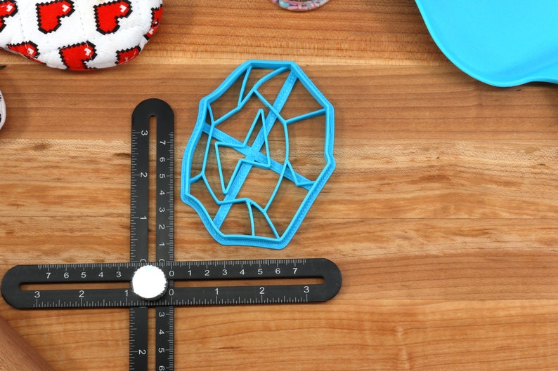 May include: A blue plastic cookie cutter shaped like a geometric design with a web-like pattern. The cutter is on a wooden surface with a ruler.