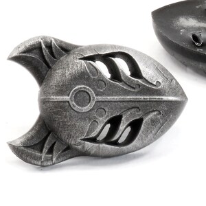 Op de afbeelding: Een zilverkleurige metalen pin in de vorm van een gestileerd vossenmasker. Het masker heeft een gedetailleerd ontwerp met ingewikkelde uitsnijdingen en een cirkelvormig patroon in het midden.
