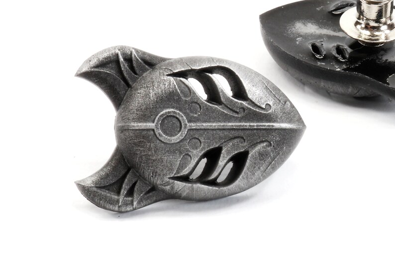 Op de afbeelding: Een zilverkleurige metalen pin in de vorm van een gestileerd vossenmasker. Het masker heeft een gedetailleerd ontwerp met ingewikkelde uitsnijdingen en een cirkelvormig patroon in het midden.