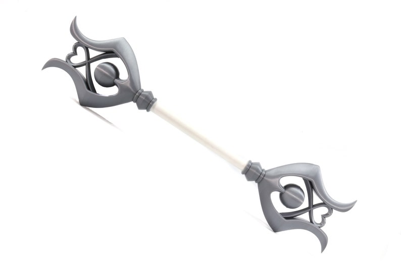May include: A silver-colored scepter with a decorative, ornate design featuring hearts and circles. The scepter has a white shaft.