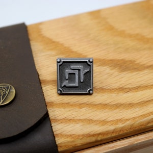 May include: A silver metal pin with a square design featuring a stylized letter "R" inside. The pin is on a wooden surface.