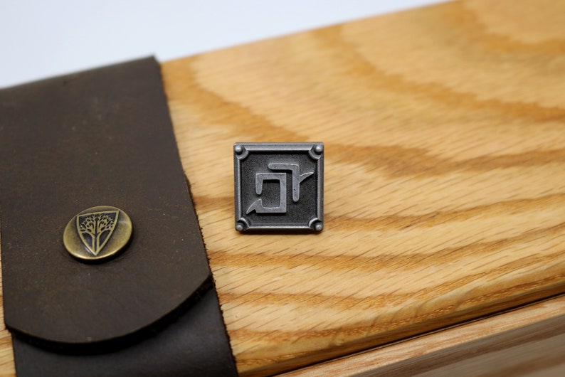 May include: A silver metal pin with a square design featuring a stylized letter "R" inside. The pin is on a wooden surface.