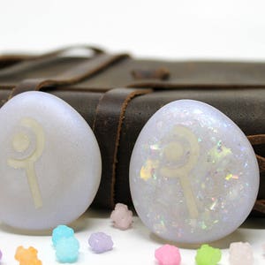 K&ouml;nnte beinhalten: Zwei wei&szlig;e, irisierende Harz-Cabochons mit einem wei&szlig;en Symbol eines Zauberstabs und einer Mondsichel auf einem braunen Lederhintergrund.