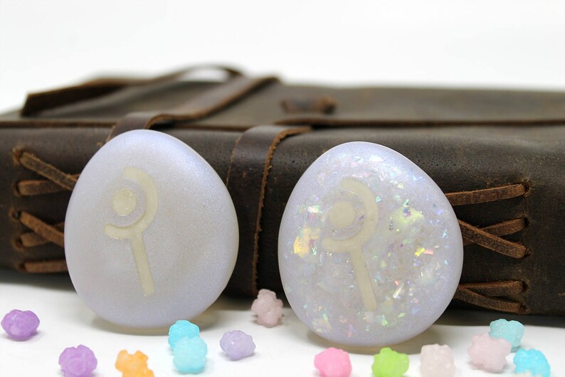 K&ouml;nnte beinhalten: Zwei wei&szlig;e, irisierende Harz-Cabochons mit einem wei&szlig;en Symbol eines Zauberstabs und einer Mondsichel auf einem braunen Lederhintergrund.