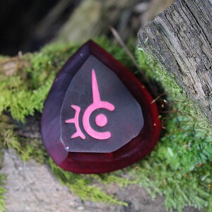 May include: A dark red, teardrop-shaped crystal with a pink symbol on the front. The symbol is a circle with a point at the top and a smaller circle inside. The crystal is sitting on a mossy log.