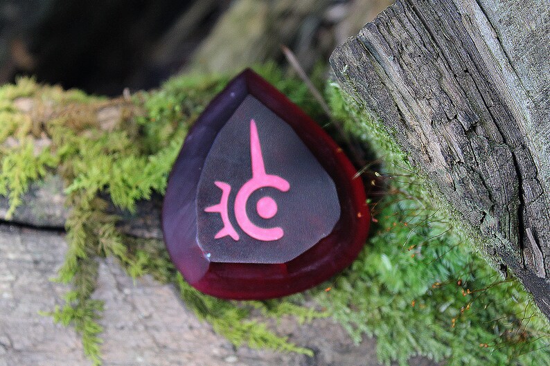 May include: A dark red, teardrop-shaped crystal with a pink symbol on the front. The symbol is a circle with a point at the top and a smaller circle inside. The crystal is sitting on a mossy log.
