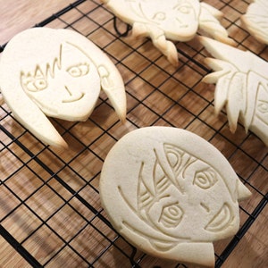 May include: Four sugar cookies decorated with icing and images of anime characters. The cookies are on a black wire cooling rack.