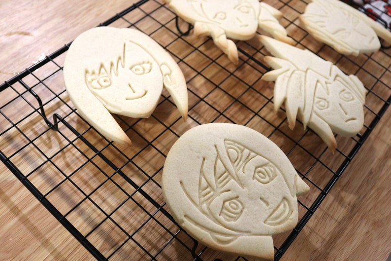 May include: Four sugar cookies decorated with icing and images of anime characters. The cookies are on a black wire cooling rack.