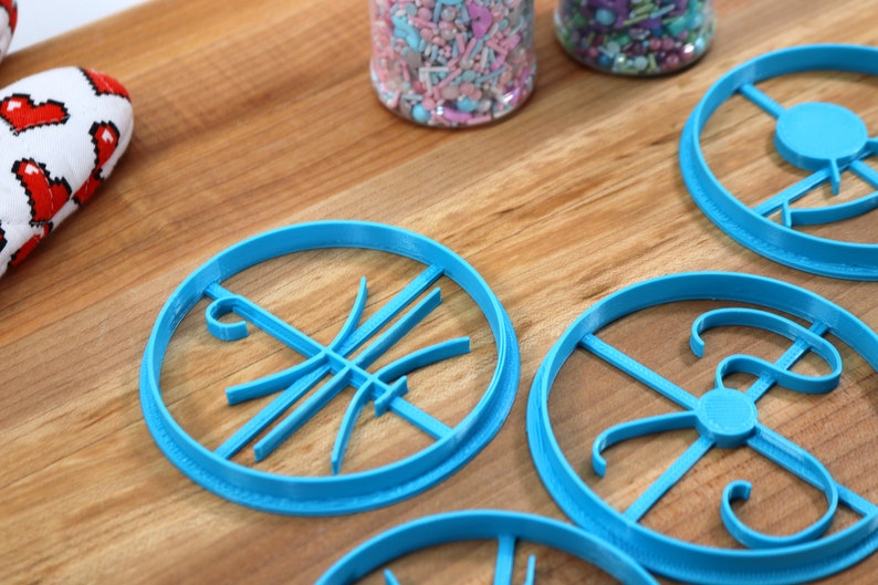 Op de afbeelding: Vier blauwe plastic koekjesvormen op een houten oppervlak. De vormen zijn in verschillende ontwerpen, waaronder een basketbal, een letter en een werveling.