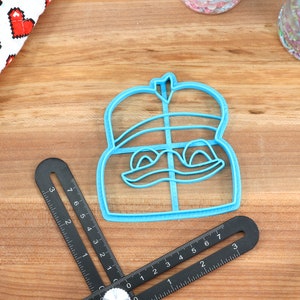 Puede incluir: Un cortador de galletas de pl&aacute;stico azul con forma de personaje de dibujos animados con una corona en la cabeza. El personaje tiene una amplia sonrisa y ojos grandes.