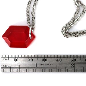 May include: A red, faceted, hexagonal pendant hangs from a silver chain. The pendant is about 2 cm wide and is measured against a ruler.