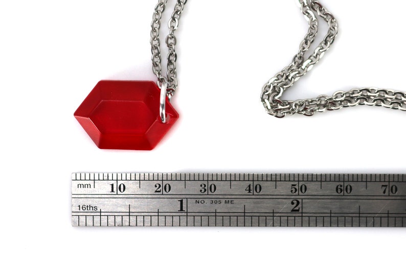 May include: A red, faceted, hexagonal pendant hangs from a silver chain. The pendant is about 2 cm wide and is measured against a ruler.
