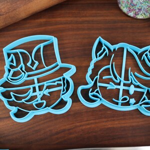 May include: Two blue plastic cookie cutters, one shaped like a person wearing a hat and the other shaped like a person with a ponytail.