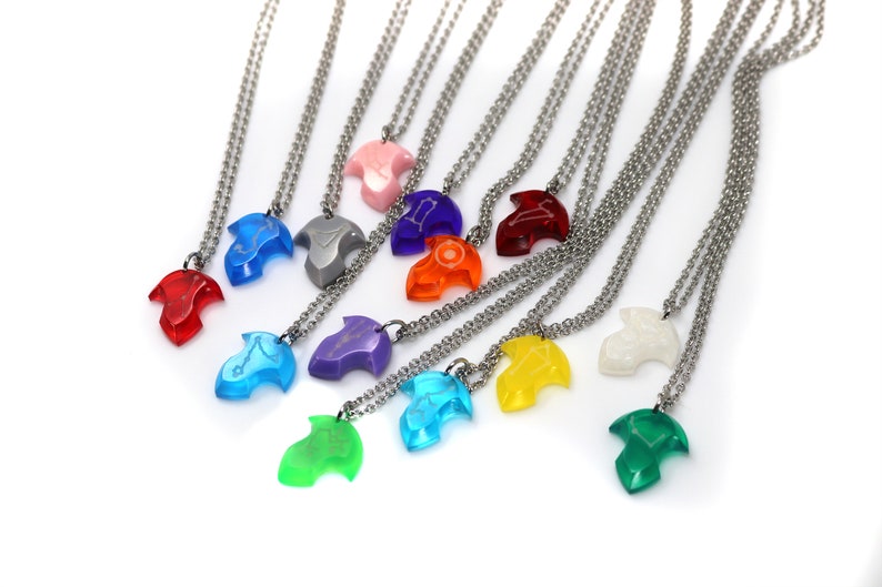Puede incluir: Un conjunto de 12 collares con colgantes de diferentes colores. Los colgantes tienen forma de flecha estilizada y tienen una cadena plateada. Los colores de los colgantes son rojo, azul, verde, morado, amarillo, naranja, rosa, blanco, gris y azul claro.