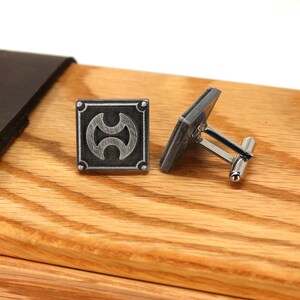May include: A pair of silver cufflinks with a geometric design on a wooden surface. The cufflinks are in a box with a leather lid.