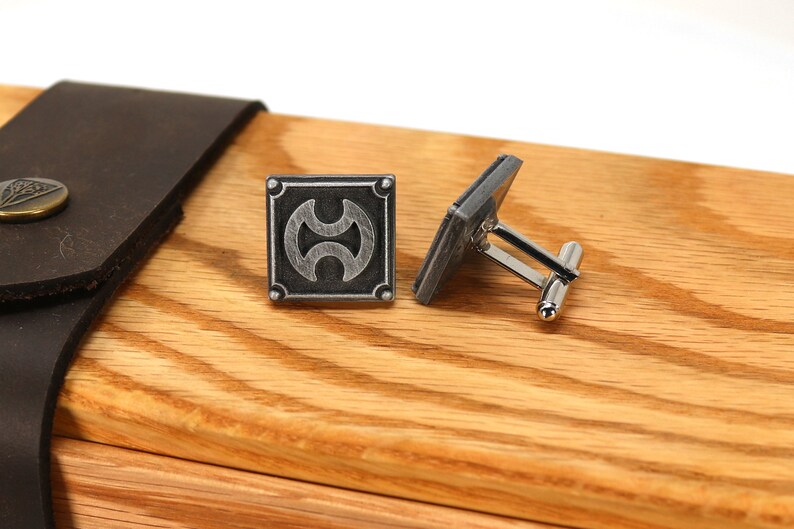 May include: A pair of silver cufflinks with a geometric design on a wooden surface. The cufflinks are in a box with a leather lid.