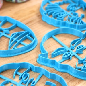 May include: Four blue plastic cookie cutters shaped like faces. The cutters are on a wooden surface.