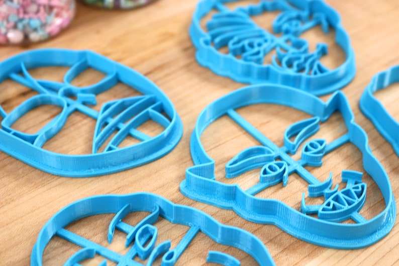 May include: Four blue plastic cookie cutters shaped like faces. The cutters are on a wooden surface.