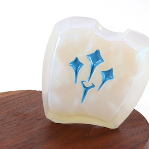 Pu&ograve; includere: Un oggetto bianco, iridescente, a forma di scudo con un design blu stilizzato di tre stelle. L'oggetto &egrave; posto su una base di legno.