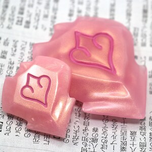 May include: Two pink, heart-shaped decorative objects with a raised heart design in a darker pink. The objects have a pearlescent finish and are resting on a newspaper background.