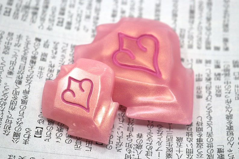 May include: Two pink, heart-shaped decorative objects with a raised heart design in a darker pink. The objects have a pearlescent finish and are resting on a newspaper background.