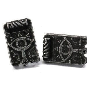 May include: A pair of silver-toned metal earrings shaped like a closed book with a stylized eye and triangle design.