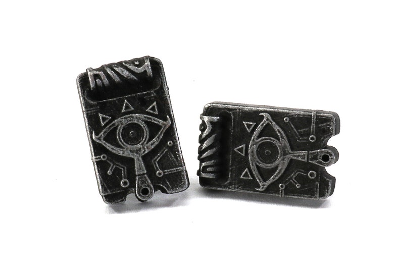 May include: A pair of silver-toned metal earrings shaped like a closed book with a stylized eye and triangle design.