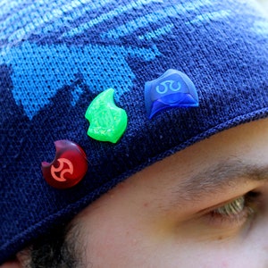 May include: A blue knitted beanie with three colourful plastic pins attached to it. The pins are shaped like a crescent moon and have different designs.