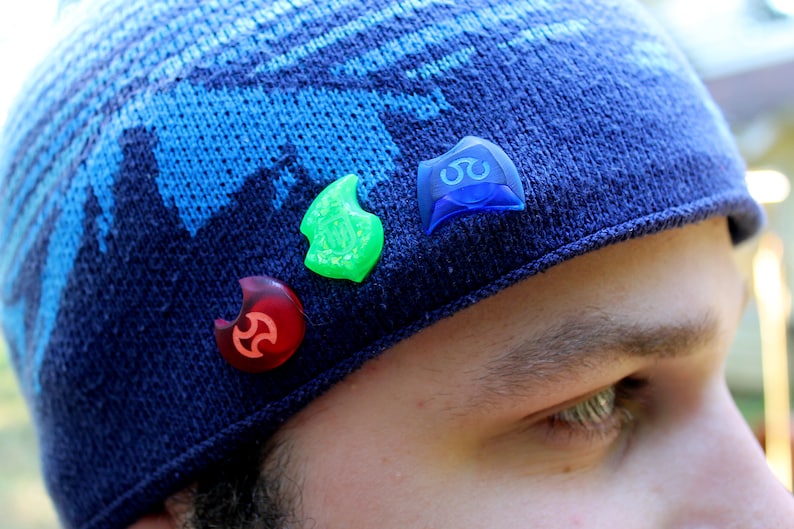 May include: A blue knitted beanie with three colourful plastic pins attached to it. The pins are shaped like a crescent moon and have different designs.