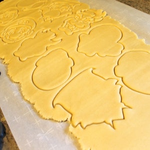 May include: A sheet of yellow cookie dough with various shapes cut out using cookie cutters. The shapes include a cloud, a tree, and a face.