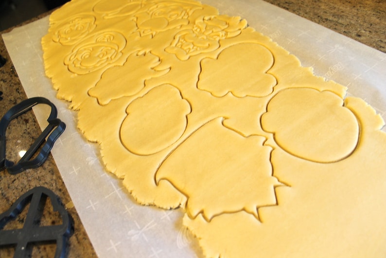 May include: A sheet of yellow cookie dough with various shapes cut out using cookie cutters. The shapes include a cloud, a tree, and a face.