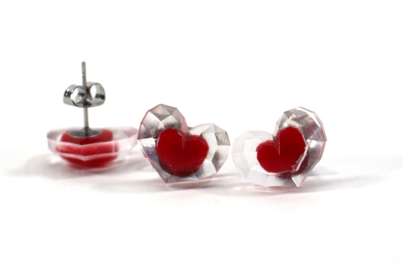 Puede incluir: Un par de pendientes transparentes en forma de coraz&oacute;n con corazones rojos en su interior. Los pendientes tienen postes de plata.