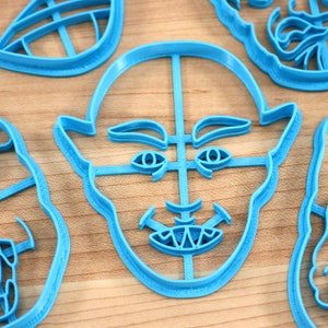 May include: Four blue plastic cookie cutters shaped like different faces. The cutters are on a wooden surface.