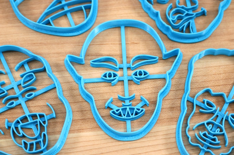 May include: Four blue plastic cookie cutters shaped like different faces. The cutters are on a wooden surface.