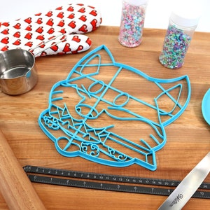 May include: A blue plastic cat-shaped cookie cutter on a wooden surface with a rolling pin, a measuring cup, a spatula, and two jars of sprinkles.