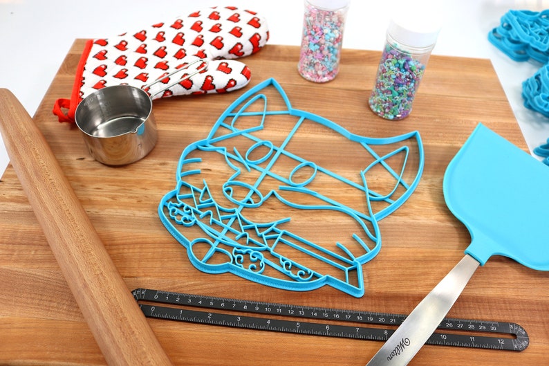 May include: A blue plastic cat-shaped cookie cutter on a wooden surface with a rolling pin, a measuring cup, a spatula, and two jars of sprinkles.