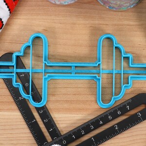 May include: A blue plastic cookie cutter shaped like a dumbbell. The cutter is on a wooden surface with a black ruler.