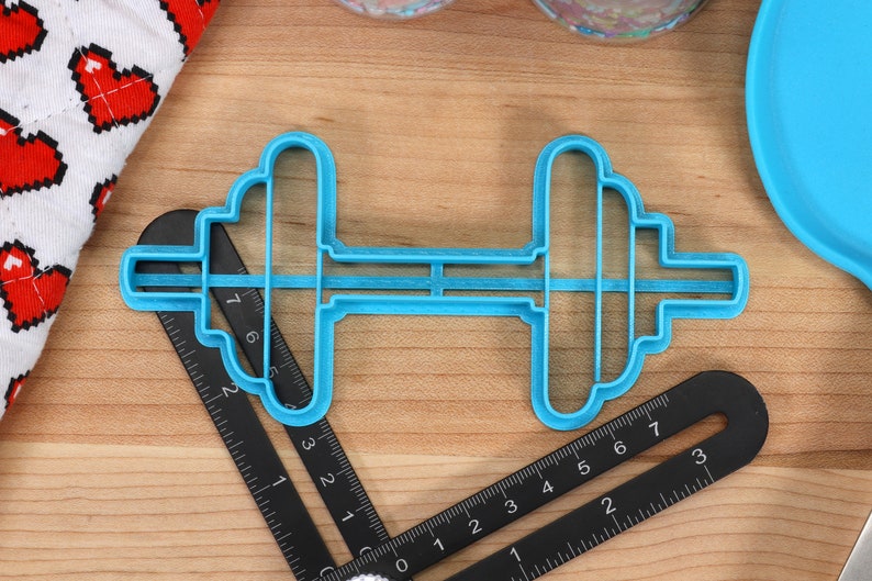 May include: A blue plastic cookie cutter shaped like a dumbbell. The cutter is on a wooden surface with a black ruler.