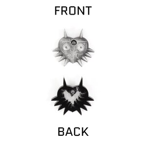 May include: A silver-colored Majora's Mask pendant with a black and white image of the mask on the back. The front of the pendant features a detailed design of the mask with a hole in the center.
