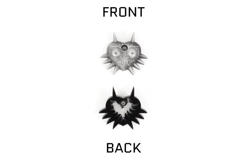 May include: A silver-colored Majora's Mask pendant with a black and white image of the mask on the back. The front of the pendant features a detailed design of the mask with a hole in the center.