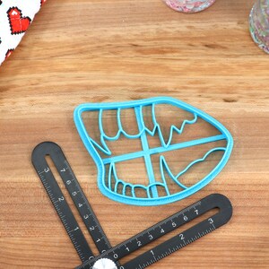 May include: A blue plastic cookie cutter shaped like a mouth with teeth. The cutter is on a wooden surface.