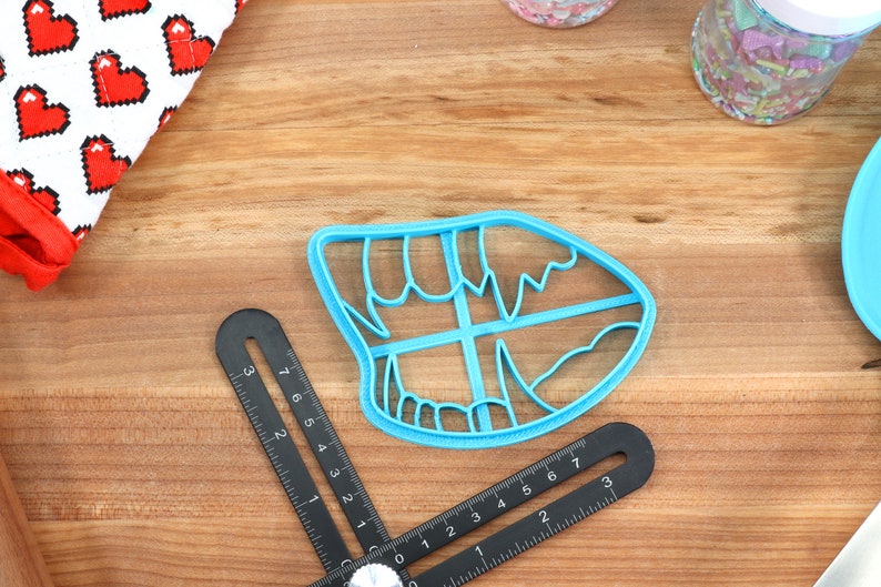 May include: A blue plastic cookie cutter shaped like a mouth with teeth. The cutter is on a wooden surface.