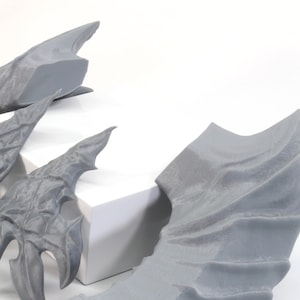 Pode incluir: Um conjunto de cinco asas de drag&atilde;o cinzas impressas em 3D. As asas s&atilde;o de v&aacute;rios tamanhos e formas, algumas t&ecirc;m uma textura mais detalhada do que outras.