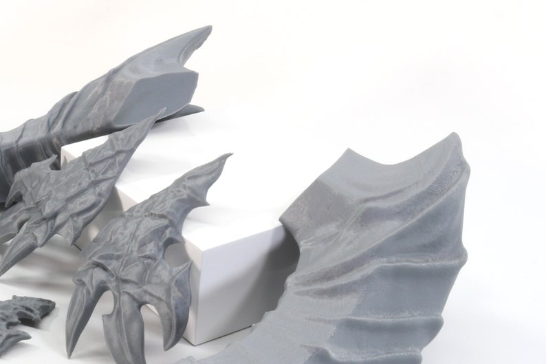 Pode incluir: Um conjunto de cinco asas de drag&atilde;o cinzas impressas em 3D. As asas s&atilde;o de v&aacute;rios tamanhos e formas, algumas t&ecirc;m uma textura mais detalhada do que outras.