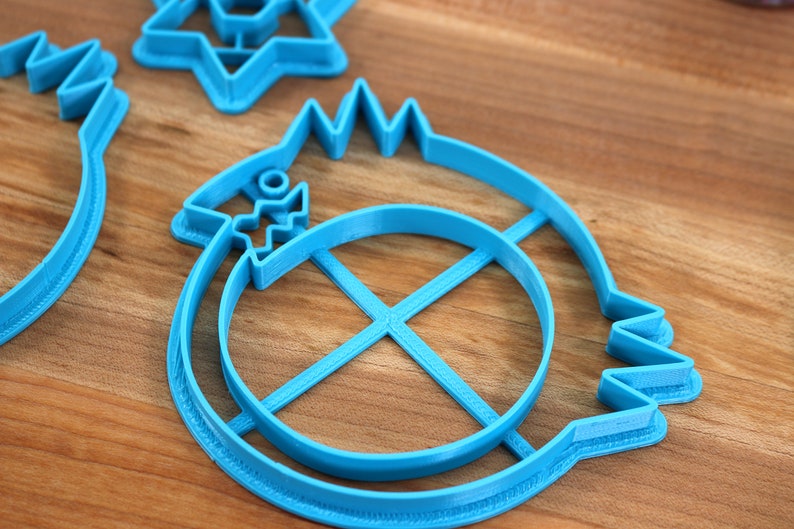 May include: A set of three blue plastic cookie cutters. The cutters are shaped like a star, a circle with a cross inside, and a fish with a circle inside.