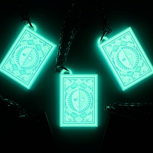 Puede incluir: Tres collares que brillan en la oscuridad con un colgante rectangular que presenta un dise&ntilde;o blanco y negro de una cara con un sol y una luna a cada lado. Los colgantes est&aacute;n en cadenas negras.