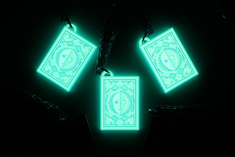Puede incluir: Tres collares que brillan en la oscuridad con un colgante rectangular que presenta un dise&ntilde;o blanco y negro de una cara con un sol y una luna a cada lado. Los colgantes est&aacute;n en cadenas negras.