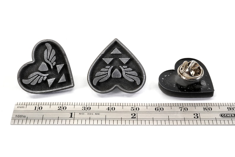 Puede incluir: Tres pines de coraz&oacute;n plateados con diferentes dise&ntilde;os. El primer pin presenta un s&iacute;mbolo alado con un dise&ntilde;o triangular. El segundo pin presenta un s&iacute;mbolo alado con un dise&ntilde;o triangular. El tercer pin es negro con un respaldo de pin plateado.