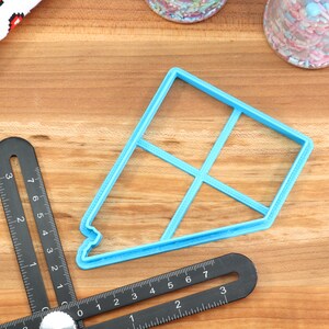 May include: A light blue plastic cookie cutter shaped like a house with a grid pattern. The cutter is on a wooden surface with a ruler and other baking supplies.